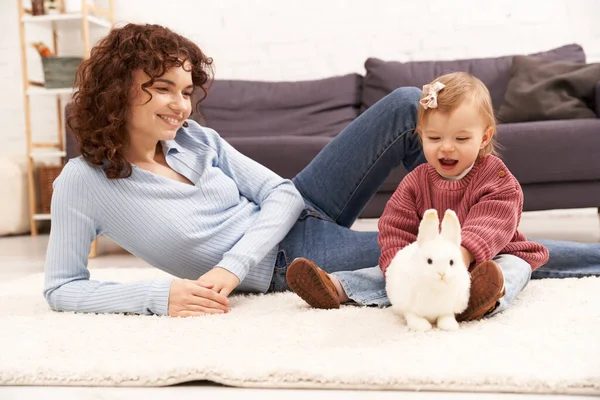 Взаимодействие с ребенком, счастливая и кудрявая женщина сидит на ковре с дочкой малыша в уютной гостиной, играя с кроликом, качество семейного времени, случайный наряд, связи между матерью и ребенком — стоковое фото