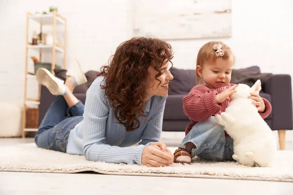 Engaging with kid, happy woman lying on carpet with toddler daughter in cozy living room, playing with rabbit, quality family time, casual attire, bonding between mother and child, motherhood — Stock Photo