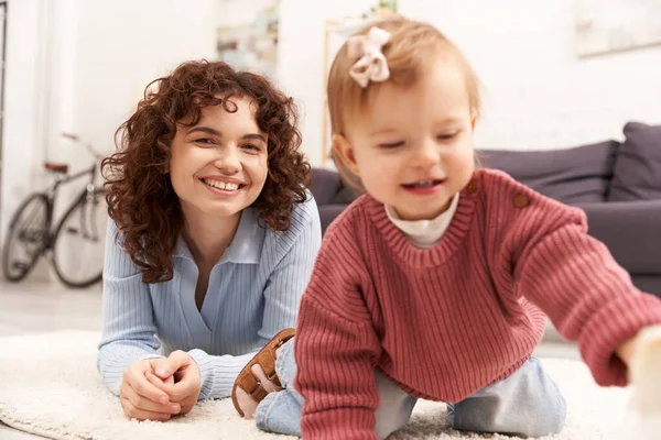 Engaging with kid, happy and curly woman lying on carpet with toddler daughter in cozy living room, quality family time, casual attire, bonding between mother and child — Stock Photo