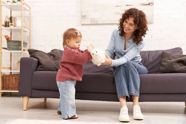 Engaging with kid, happy and curly woman sitting on couch with toddler daughter in cozy living room, playing with rabbit, quality family time, casual attire, bonding between mother and child — Stock Photo