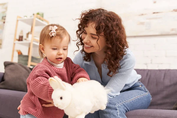 Work life balance, positive and curly woman sitting on couch with toddler daughter in cozy living room, playing with rabbit, quality family time, casual attire, bonding between mother and child — Stock Photo