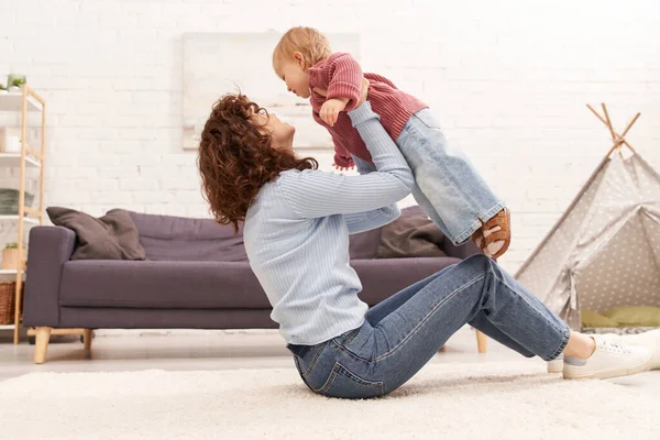 Quality family time, curly woman lifting toddler daughter and sitting on carpet in cozy living room, work life balance, denim clothes, casual attire, family relationships, modern parenting — Stock Photo