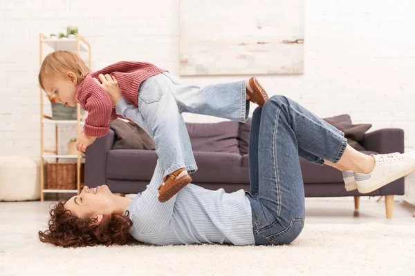 Качественное семейное время, счастливая мать поднимая малыша дочь и лежа на ковре в уютной гостиной, баланс трудовой жизни, джинсовая одежда, случайный наряд, семейные отношения, современное воспитание — стоковое фото