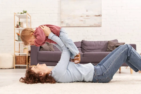 Pais modernos mãe feliz levantando filha da criança e deitado no tapete na acolhedora sala de estar, equilíbrio de vida no trabalho, roupas jeans, traje casual, relacionamentos familiares, tempo de qualidade — Fotografia de Stock