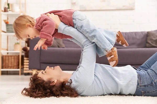 Современное воспитание счастливая мать поднимая малыша дочь и лежа на ковре в уютной гостиной, баланс трудовой жизни, джинсовой одежды, случайный наряд, привлечение с ребенком, качество времени — стоковое фото