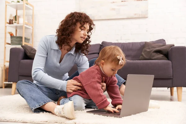 Modern working parent, engaging with child, balancing work and life, curly woman supporting baby girl near laptop, modern parenting, multitasking woman, freelance, building successful career — Stock Photo