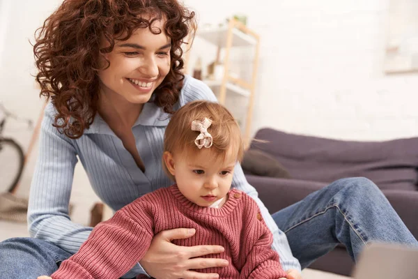 Multitasking woman, engaging with child, work and life balance, curly woman supporting toddler baby girl in cozy living room, modern parenting, loving motherhood, positivity — Stock Photo