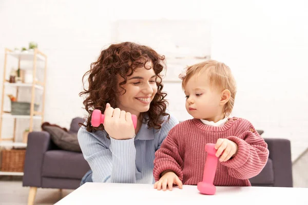 Time management, working mother, balanced lifestyle, curly woman exercising with dumbbell near toddler daughter in cozy living room, home workout, sport, busy mom, modern parenting — Stock Photo