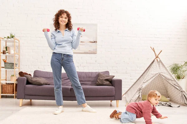 Time management, working mother, balanced lifestyle, happy woman exercising with dumbbells near toddler daughter in cozy living room, home workout, sport, busy mom, physical activity, interior — Stock Photo