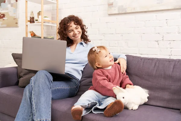 Multitasking woman, freelance, curly woman using laptop and sitting on couch with baby girl in cozy living room, modern parenting, building successful career, balancing work and life — Stock Photo