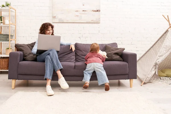 Багатозадачна жінка, позаштатна, кучерява жінка використовує ноутбук і сидить на дивані біля дочки малюка в затишній вітальні, сучасне батьківство, будуючи успішну кар'єру, балансуючи роботу і життя — стокове фото