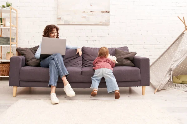 Mujer multitarea, independiente, mujer rizada utilizando el ordenador portátil y sentado en el sofá cerca de la hija del niño en la acogedora sala de estar, la crianza moderna, la construcción de una carrera exitosa, equilibrar el trabajo y la vida - foto de stock