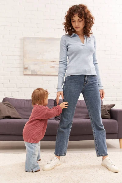 Повнометражний малюк робить свої перші кроки поблизу кучерявої матері, затишної вітальні, займається дитиною, джинсових джинсів, повсякденного одягу, сімейного часу, сучасного батьківства, життєвого балансу — стокове фото