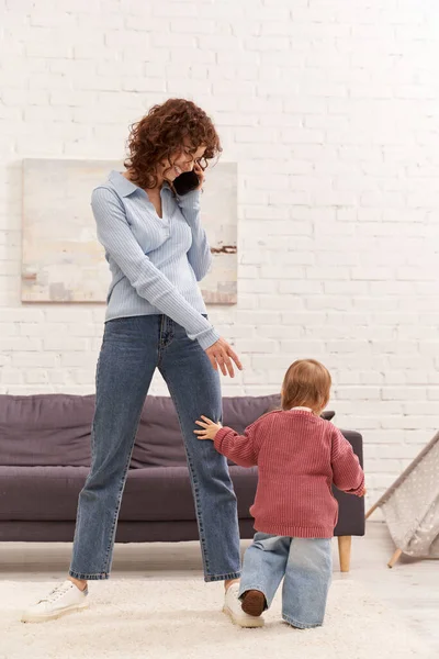 Full length of toddler baby doing her first steps near happy mother, cozy living room, engaging with kid, denim jeans, casual attire, family time, modern parenting, work life balance — Stock Photo