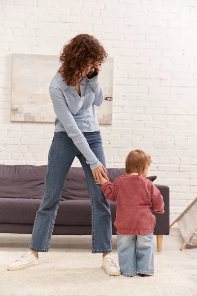 Comprimento total do bebê da criança fazendo seus primeiros passos perto da mãe encaracolada, engajando-se com a criança, jeans jeans jeans, traje casual, tempo da família, paternidade moderna, equilíbrio da vida profissional, sala de estar aconchegante — Fotografia de Stock