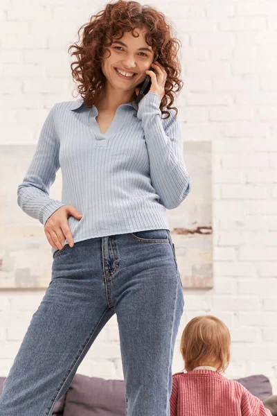 Cheerful young woman talking on smartphone standing near toddler kid, balancing between work and life, modern parenting, remote work, career and family, casual attire — Stock Photo