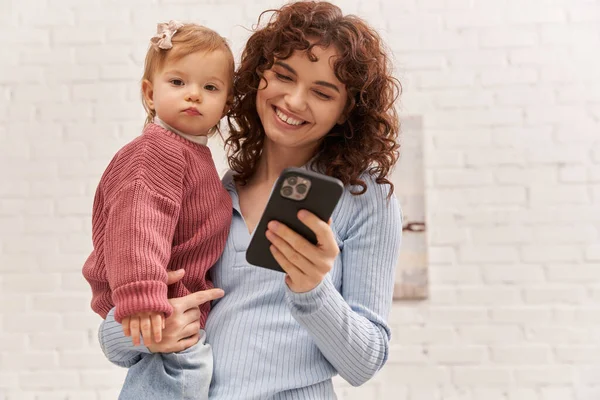 Happy young woman holding in arms baby girl and using smartphone, balancing between work and life, modern parenting, relationships, mother daughter bond, remote work, career and family — Stock Photo