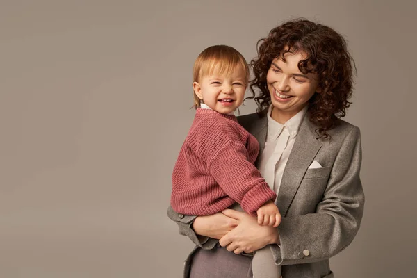 Karriere und Familie, fröhliche junge Frau mit Kleinkind im Arm, Spagat zwischen Arbeit und Leben, moderne Elternschaft, Geschäftsfrau, Mutter-Tochter-Bindung, Lebensstil, grauer Hintergrund — Stockfoto