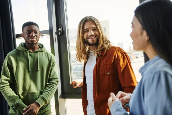 Creative multicultural business people talking and planning startup project in modern office space, long haired and stylish man smiling near african american colleague and asian woman — Stock Photo