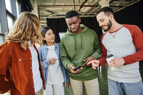 African american man in trendy casual clothes standing with mobile phone near young and multiethnic partners in modern office, creative and diverse team, business collaboration and partnership — Stock Photo