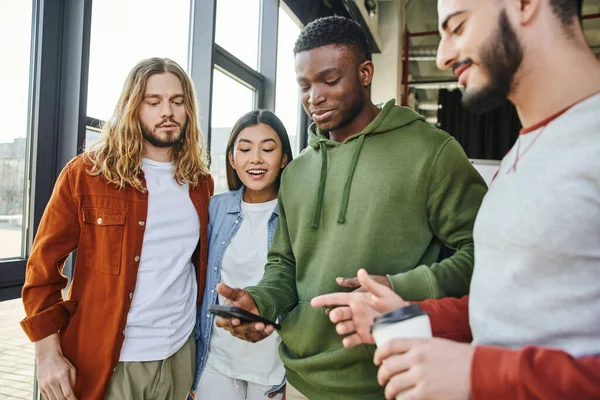 Smiling bearded man with takeaway coffee pointing at smartphone in hand of african american friend near positive multiethnic colleagues in modern office, diverse team collaboration, business startup — Stock Photo