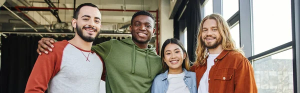 Jovem e alegre homem afro-americano abraçando amigos elegantes e ambiciosos e sorrindo para a câmera no escritório moderno, parceiros de negócios multiétnicos, startup de sucesso, banner — Fotografia de Stock