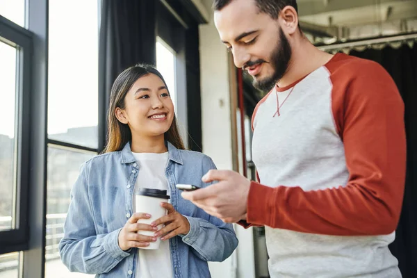 Радостная азиатская женщина в стильной повседневной одежде держит кофе, чтобы пойти и глядя на бородатый мужчина просматривает интернет на мобильном телефоне в современном офисе, молодые предприниматели, успешное сотрудничество — стоковое фото