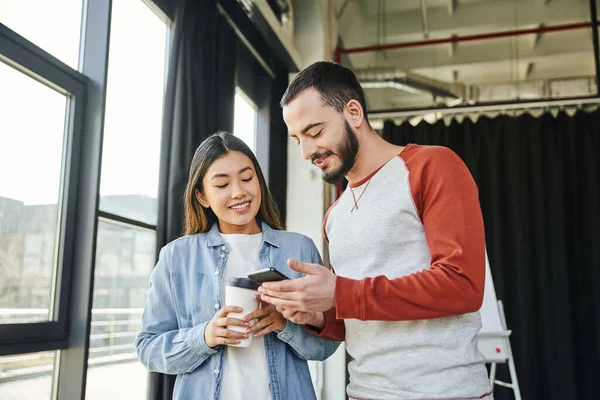 Happy bearded man showing mobile phone to attractive asian woman holding coffee to go in office, interracial business colleagues, young entrepreneurs, startup project collaboration — Stock Photo