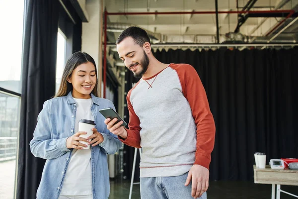 Glad and bearded man networking on smartphone near smiling asian woman with takeaway drink, youthful and creative multiethnic business colleagues in modern office space — Stock Photo