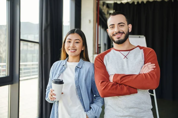 Pleased asian woman with coffee to go in paper cup and bearded man with folded arms smiling at camera in modern office, young interracial entrepreneurs collaborating in startup — Stock Photo