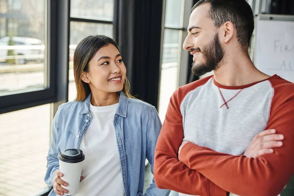 Young and successful interracial colleagues smiling at each other in contemporary office, asian woman with takeaway coffee and bearded man with folded arms, business startup collaboration — Stock Photo