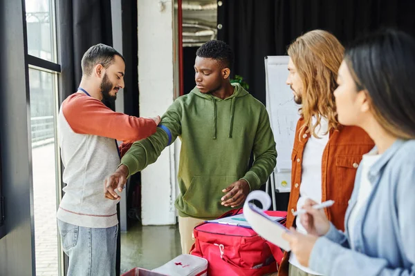 First aid training seminar, bearded medical instructor applying compression tourniquet on arm of african american man near young participants with clipboards, safety and emergency response concept — Stock Photo