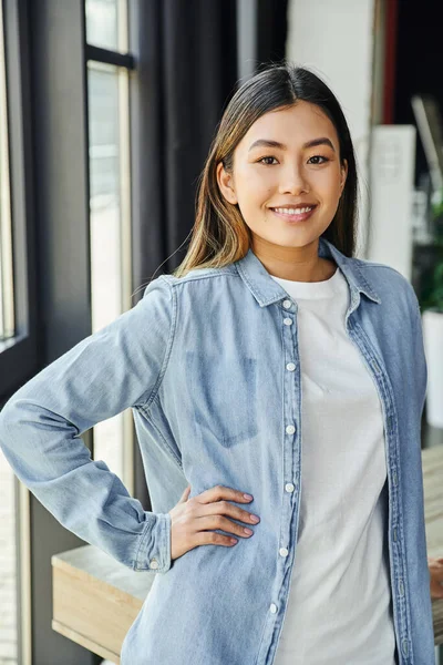 Jovem e atraente asiática empresária em camisa jeans azul, com cabelo morena e sorriso radiante em pé com a mão no quadril e olhando para a câmera no escritório moderno, emoção positiva — Fotografia de Stock