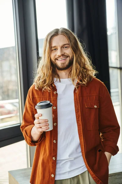 Homem hipster overjoyed segurando takeaway café em copo de papel, de pé com a mão no bolso e olhando para a câmera no escritório moderno, cabelo longo, barba, camisa da moda, empreendedor ambicioso — Fotografia de Stock