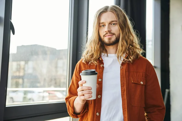 Gutaussehender und cooler Geschäftsmann mit Pappbecher mit Kaffee zum Mitnehmen, während er am Fenster steht und im Büro in die Kamera schaut, lange blonde Haare, trendiges Hemd, Hipster-Stil — Stockfoto