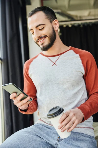 Charismatic entrepreneur, bearded and stylish, holding coffee to go in paper cup and browsing internet on smartphone, young and career oriented businessman in modern office — Stock Photo