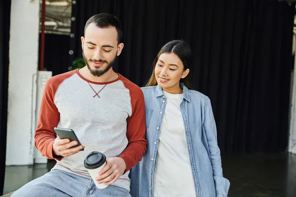 Homem barbudo positivo segurando copo de papel com café para ir e navegar na internet no smartphone perto de sorrir mulher asiática, colegas inter-raciais, colaboração empresarial bem sucedida — Fotografia de Stock