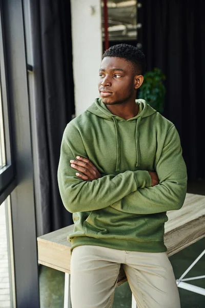 Задумчивый африканский американец в зеленой стильной толстовке сидит на столе со сложенными руками и смотрит в сторону в современном офисе, молодой бизнесмен, профессиональное развитие — стоковое фото