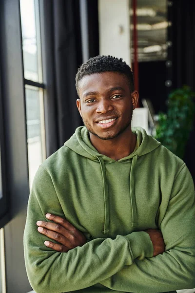 Jovem e carismático empresário afro-americano em verde e moderno capuz de pé com braços dobrados e sorrindo para a câmera, emoção feliz, espaço de escritório contemporâneo — Fotografia de Stock