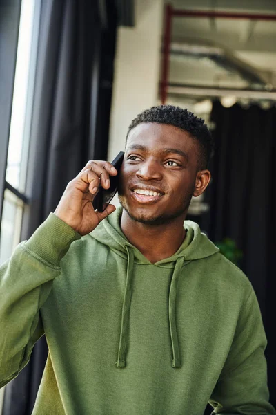 Optimistic african american man in green hoodie talking on mobile phone and looking away in contemporary office, successful entrepreneur discussing business startup — Stock Photo