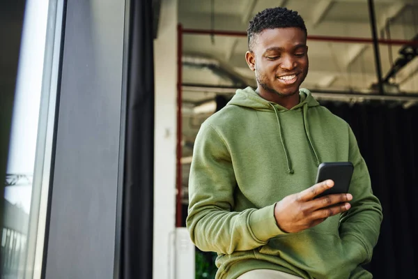 Jeune homme afro-américain joyeux tenant la main dans la poche de sweat à capuche vert et naviguant sur Internet sur téléphone portable dans un bureau moderne, homme d'affaires prospère, émotion positive — Photo de stock
