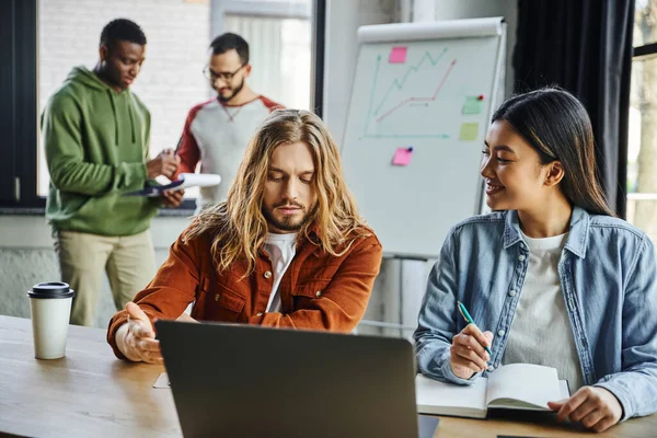 Long haired man pointing at laptop near smiling asian woman while interracial colleagues working near flip chart with graphs on blurred background, multicultural collaboration in modern office — Stock Photo