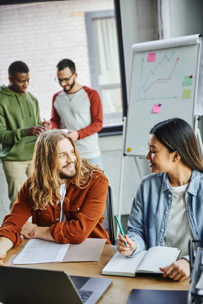 Positive long haired man and young asian woman talking near notebook, documents and laptop while interracial colleagues working at flip chart with graphs on blurred background in modern office — Stock Photo