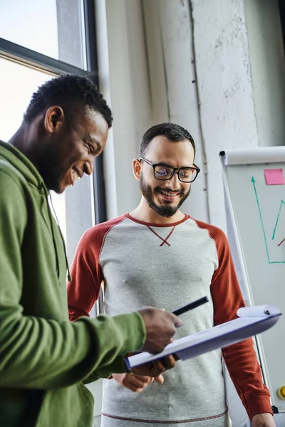 Young and cheerful african american entrepreneur showing documents on clipboard to african american business partner in eyeglasses while working on startup in office, successful collaboration — Stock Photo