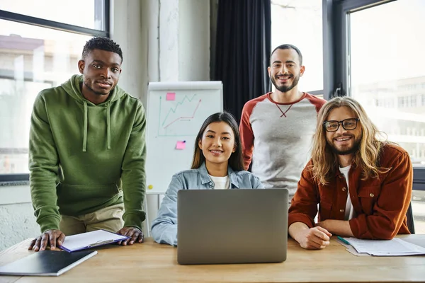 Stylish and ambitious multicultural colleagues smiling at camera near laptop and flip chart with graphs on blurred background, collaboration of young and successful entrepreneurs — Stock Photo