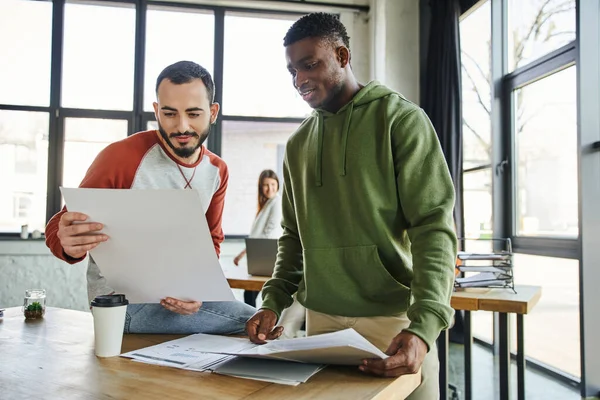 Young and ambitious business partners working on startup planning in modern office, bearded man holding document near african american colleague, creative thinking and collaboration — Stock Photo