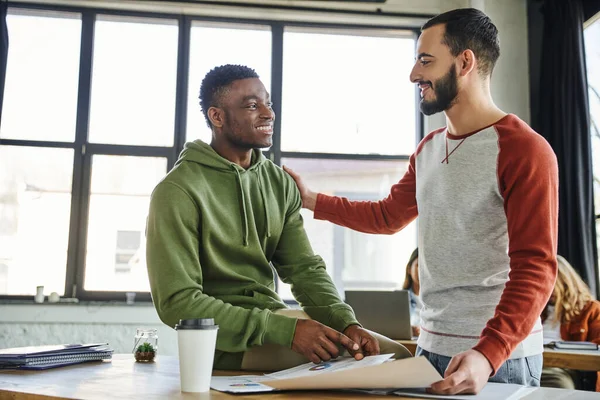 Homem barbudo satisfeito com colega afro-americano sorrindo uns para os outros perto de documentos e xícara de papel com café no escritório moderno, colaboração bem sucedida de jovens empresários inter-raciais — Fotografia de Stock
