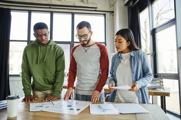 Empreendedores multiculturais em roupas casuais olhando para documentos com gráficos em mesa no escritório moderno, equipe jovem e criativa trabalhando no planejamento de startup, parceria bem sucedida — Fotografia de Stock