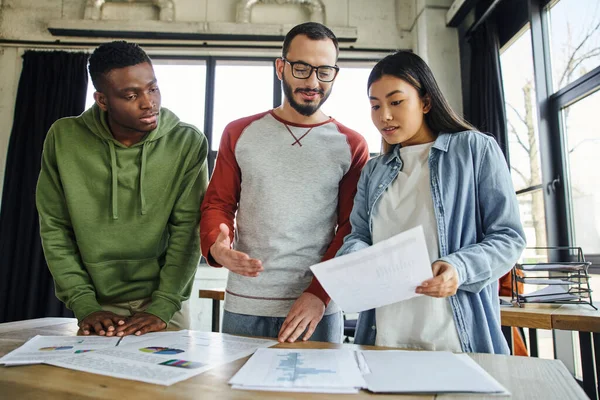 Young asian businesswoman showing document to bearded man in eyeglasses and african american colleague while working on startup planning in contemporary office — Stock Photo