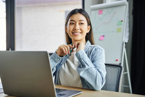 Веселая и мечтательная азиатская бизнесвумен в синей джинсовой рубашке сидит перед ноутбуком, держит ручку и смотрит в сторону от флип-чарта на размытом фоне в современном офисе — стоковое фото